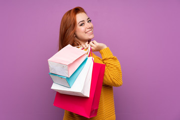 Redhead teenager girl over isolated purple background holding shopping bags and smiling