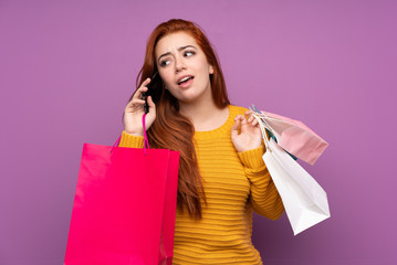 Redhead teenager girl over isolated purple background holding shopping bags and calling a friend with her cell phone