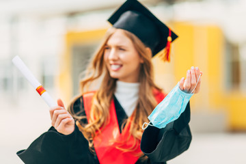 A female graduate student in a black graduation dress, holding a protective medical mask in her hand. Graduation ceremony concept, quarantine, coronavirus
