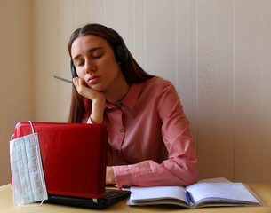 A girl with headphones is napping at the table with a computer and a copybooks. Distance education during the quarantine due to the epidemic of coronavirus Covid-19. Home schooling content