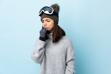 Mixed race skier girl with snowboarding glasses over isolated blue background having doubts.