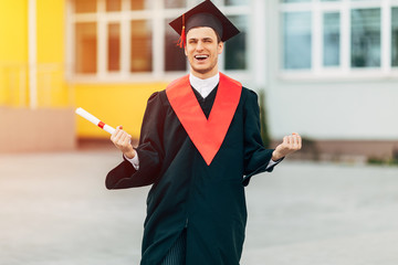 A male graduate student in a black graduation dress, holding a diploma, shows a gesture of victory...