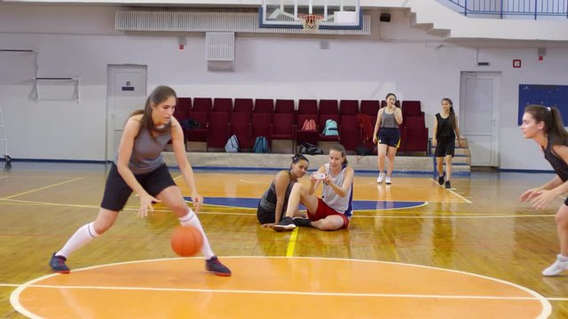 Two young female athletes in sportswear practicing dribbling and passing a basketball on court while their teammates watching their workout and recording video with smartphoneTwo young female athletes