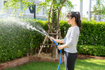 Asian woman having happy in home backyard summer garden, with garden hose splashing water on the...