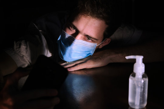 a doctor exhausted resting on the table at night quarantine lockdown for social distancing with the cellphone screen lighting , the chinstrap and the gel alcohol during the covid-19 pandemic
