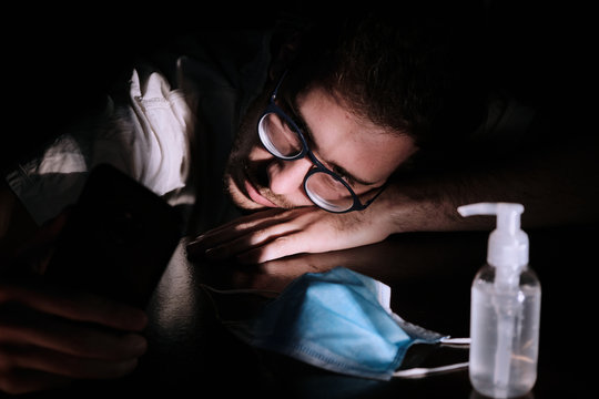 a doctor exhausted resting on the table at night quarantine lockdown for social distancing with the cellphone screen lighting , the chinstrap and the gel alcohol during the covid-19 pandemic