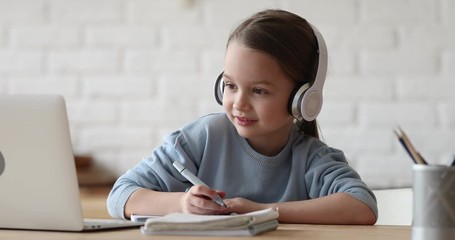 Happy small primary school girl elearning online using computer app. Child pupil wearing headphones video calling remote tutor or watching video lesson class on laptop. Kid distance study at home. - Powered by Adobe