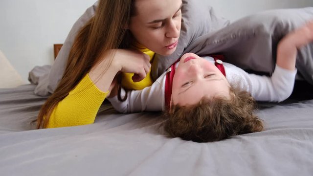 Loving happy young mother kissing cute little daughter lying on bed relish time together at home, expressing love, smiling mom and kid girl having fun playing, sincere warm relationships concept