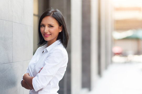Attractive Hispanic female with arms crossed and looking at camera over building background at outdoor. Smiling Beautiful woman in white shirt standing with confident expression