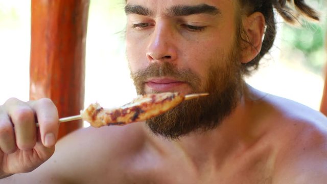 A young man with dreadlocks and a beard eats chicken skewers