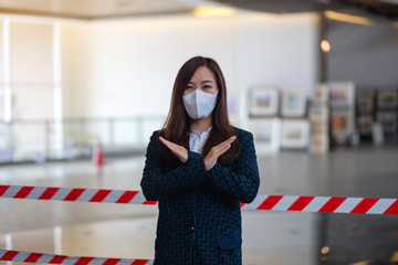 Closeup image of an asian woman wearing protective face mask, making crossed arms sign in front of red and white warning tape area for preventing the spread of Covid-19 the pandemic concept