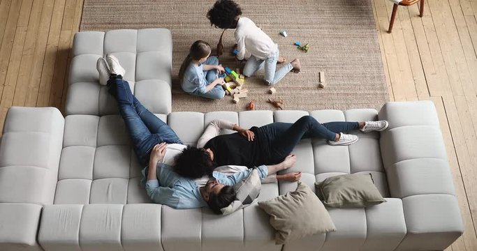 Mixed ethnicity parents relaxing on comfortable couch while cute diverse kids daughters playing on floor at home. Interracial family with two children spending time together in living room. Top view