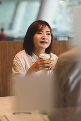 Beautiful woman relaxing and holding a take-away coffee cup and pencil in her hands while sitting at the working desk over comfortable and modern office as background.