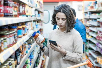 Focused woman using smartphone in grocery store. Young woman reading checklist via smartphone and...