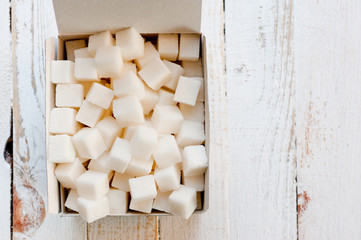 Refined sugar cubes in a cardboard box shot on a white painted wooden surface.