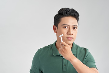 Handsome young man shaving his face and looking at camera while standing isolated on grey background