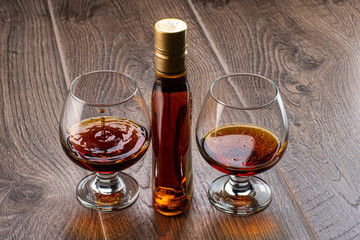 two glasses with cognac and splash next to a bottle on a wooden background