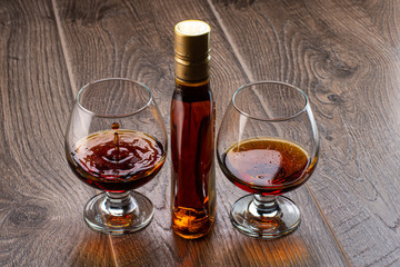 two glasses with cognac and splash next to a bottle on a wooden background