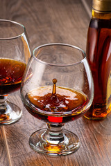 two glasses with cognac and splash next to a bottle on a wooden background