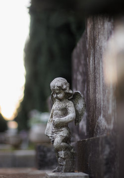 Small Sculpture Of A Baby Angel With Wings On A Stone Grave Tombstone. Old Dirty Worn Over Time