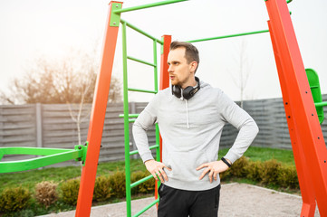 An athletic guy in hoodie and with headphones on the outdoor sports ground with a horizontal bars