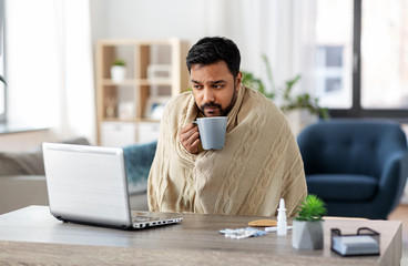 health, cold and people concept - sick young indian man in blanket with laptop computer drinking...