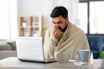 healthcare, technology and people concept - sick indian man in blanket with runny nose having video call on laptop computer at home - obrazy, fototapety, plakaty