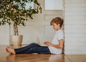 Young blond boy in white shirt and jeans printing on the white laptop sitting on the floor at home