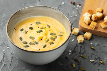 Creamy pumpkin soup with seeds and spices served with crackers on wooden boards on grey backgound. Close up view