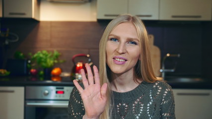 Lovely young female smiling and looking at camera while greeting vlog audience by waving hand on background of stylish kitchen.