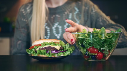 Close-up shot of Young lady preferring hamburger to salad. Attractive young woman choosing to eat healthy hamburger for breakfast while sitting at table in stylish kitchen.