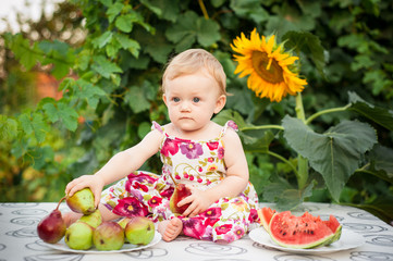 little girl of 10 months on the street in the yard in the summer in a colored dress there is a red juicy watermelon