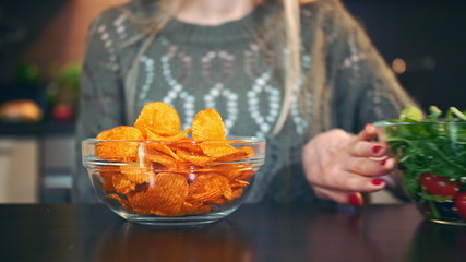 Close-up shot of lady preferring crisp to salad. Attractive young woman choosing to eat healthy crisp for lunch while sitting at table in stylish kitchen and she eats these chips.