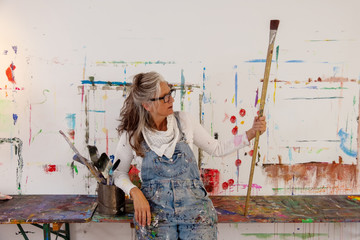 proud older artist woman, in her fifties with grey hair and black glasses holds a big paintbrush - obrazy, fototapety, plakaty