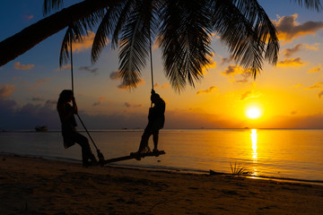 Silhouetted couple in love walks on the beach during sunset. Riding on a swing tied to a palm tree and watching the sun go down into the ocean