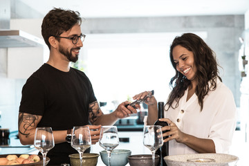 Smiling woman holding bottle of red wine and taking corkscrew. Happy young man and woman opening wine at kitchen. Wine tasting concept