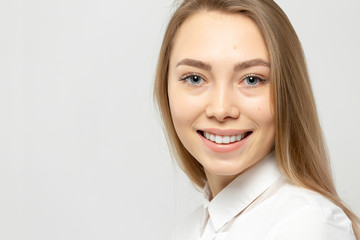 Close-up portrait of beautiful happy young attractive blonde woman in a white shirt with clean skin looking at camera against gray-white background.