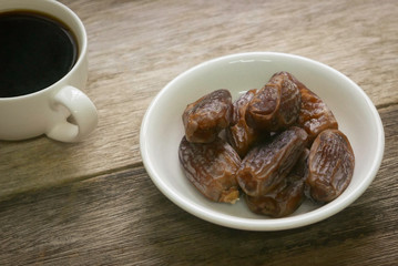 Selective focus of a dried dates fruit in a white bowl together with a cup of coffee on wooden background. Ramadan holy month for muslim concept where they are fasting.