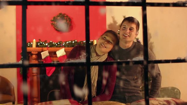 Couple looking out of window at home at Christmas with snow falling outside. Snowing. Christmas Tree and fireplace in lounge. Stock Video Clip Footage