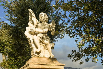Close-up of a stone statue representing a winged putto on the top of a column with tree branches and blue sky in background, Sanremo, Liguria, Italy