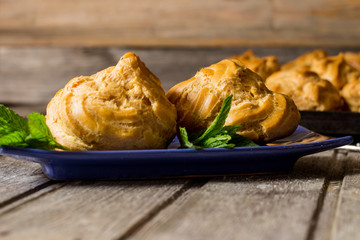 Delicious big cream puffs with cream in hand painted blue oval saucer with mint leaves on aged wooden background. Selective focus.