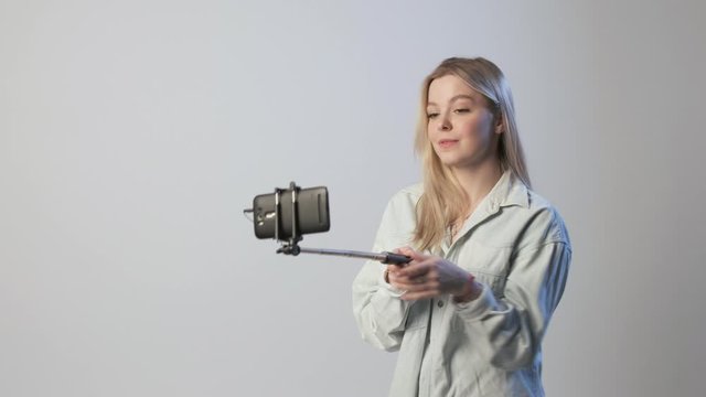 cute female records video by smartphone attached to selfie stick. blond girl filming vlog speaking, smiling, chatting with followers, using cellphone on white background. Beauty blogger recording blog