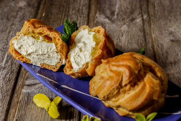 Delicious big cream puffs with cream in hand painted blue oval saucer with mint leaves on aged wooden background. Selective focus.