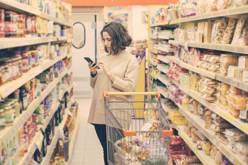 Woman with smartphone choosing goods in store. Focused young woman holding mobile phone and buying products in supermarket. Shopping concept