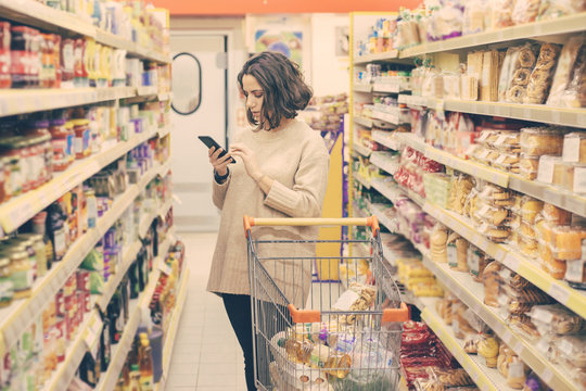 Woman with smartphone choosing goods in store. Focused young woman holding mobile phone and buying products in supermarket. Shopping concept