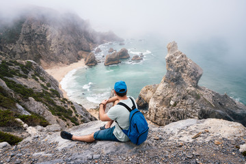 Adult male with backpack resting at stone edge of Praia da Ursa Beach coastline. Surreal scenery...