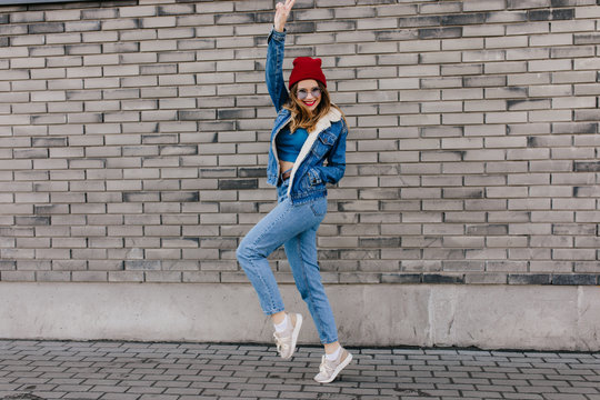 Full-length photo of shapely attractive girl funny dancing on the street. Outdoor portrait of wonderful blonde woman in red hat jumping near brick wall.