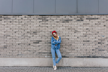 Full-length shot of gorgeous woman in white sneakers posing on urban background with arms crossed. Outdoor portrait of attractive female model in red hat standing in front of brick wall.