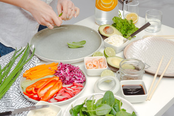 The process of cooking spring rolls - woman's hands putting basil on rice paper. Table with spring rolls ingredients and water with oranges.