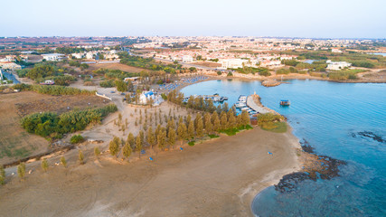 Fototapeta na wymiar Aerial bird's eye view of coastline sunset, landmark white washed chapel Agia Triada beach, Protaras, Famagusta, Cyprus from above. Ayia Trias bay church and a boat out of the small port at sunrise.